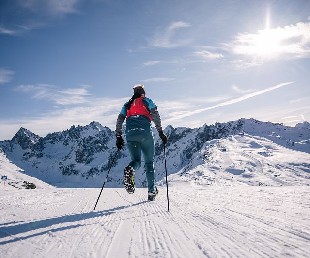 Ein Trailrunner in blauer Ausrüstung fährt bei sonnigem Wetter eine schneebedeckte Piste hinunter. Im Hintergrund sind beeindruckende, schneebedeckte Berge zu sehen.