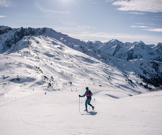 Eine Trailrunner läuft bei sonnigem Wetter auf Skiern einen verschneiten Hang hinunter. Im Hintergrund sind beeindruckende, schneebedeckte Berge und ein klarer blauer Himmel zu sehen.