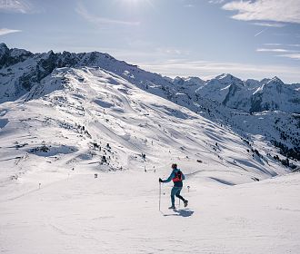 Eine Trailrunner läuft bei sonnigem Wetter auf Skiern einen verschneiten Hang hinunter. Im Hintergrund sind beeindruckende, schneebedeckte Berge und ein klarer blauer Himmel zu sehen.