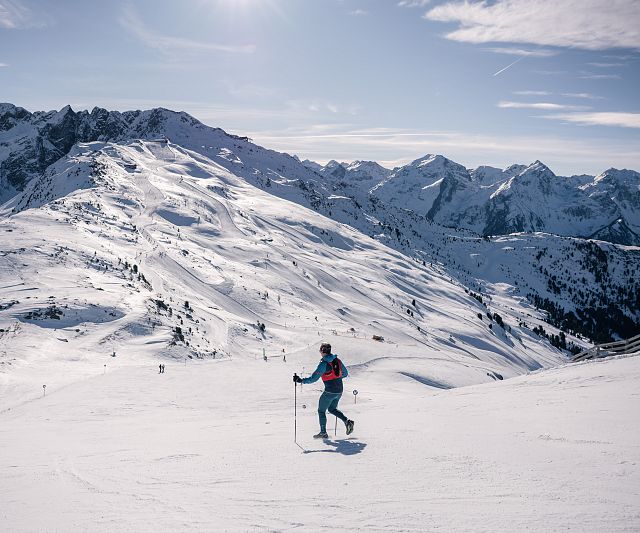 Eine Trailrunner läuft bei sonnigem Wetter auf Skiern einen verschneiten Hang hinunter. Im Hintergrund sind beeindruckende, schneebedeckte Berge und ein klarer blauer Himmel zu sehen.