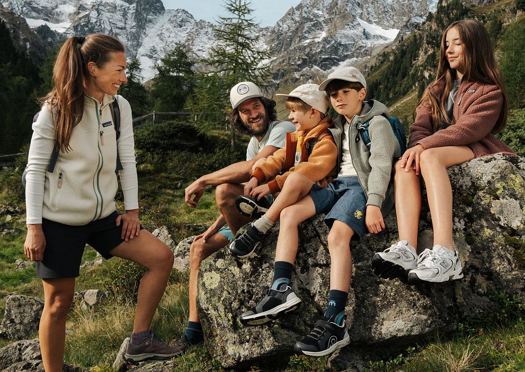 Eine Familie entspannt sich in einer alpinen Landschaft. Zwei Erwachsene und drei Kinder sitzen auf Felsen, umgeben von grünen Bäumen und schneebedeckten Bergen im Hintergrund.