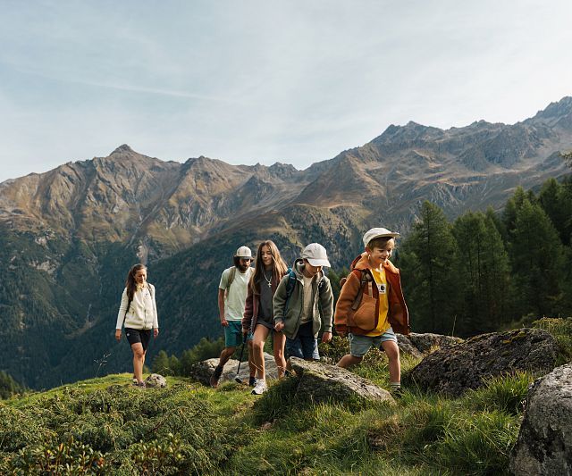 Familie mit drei Kindern wandert auf einem grasbewachsenen Hügel vor einer beeindruckenden Bergkulisse. Sie tragen Freizeitkleidung und genießen die Natur.