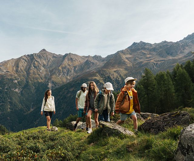Familie mit drei Kindern wandert auf einem grasbewachsenen Hügel vor einer beeindruckenden Bergkulisse. Sie tragen Freizeitkleidung und genießen die Natur.