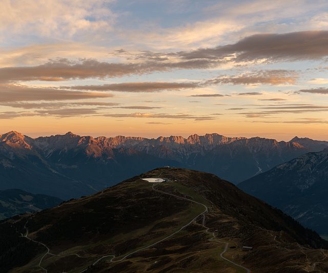 Berglandschaft bei Sonnenaufgang mit Bergen im Hintergrund. Ein kleiner Wasserteich liegt auf einem Hügel, über dem der Himmel in warmen Farbtönen leuchtet.