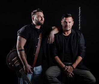 Zwei Männer posieren vor einem dunklen Hintergrund. Einer steht mit einer Gitarre, der andere sitzt. Beide tragen Freizeitkleidung und schauen ernst.