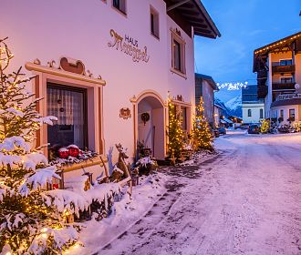 Verschneite Dorfstraße bei Dämmerung, gesäumt von Häusern mit festlicher Beleuchtung. Tannenbäume und Lichterketten schmücken die Szenerie, Berge im Hintergrund.