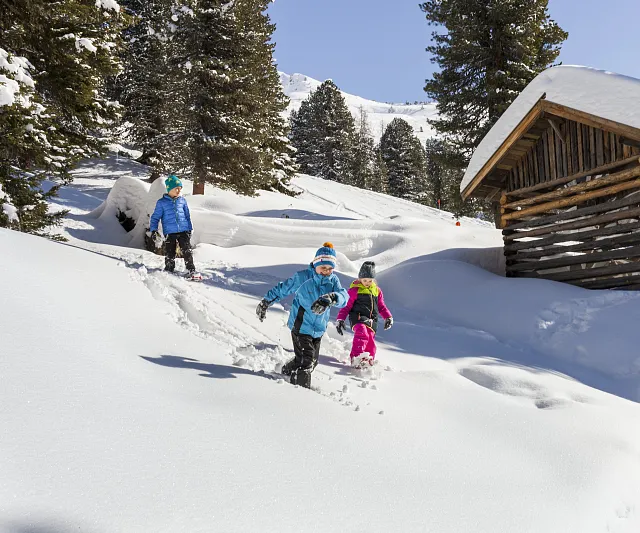 Zwei Kinder und ein Erwachsener laufen fröhlich durch den tiefen Schnee in der Nähe einer Holzhütte.