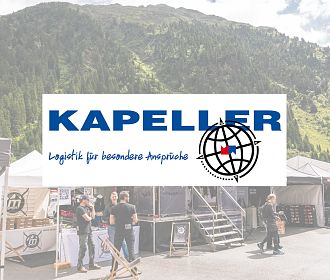 Das Bild zeigt einen Stand mit dem Logo von Kapeller. Im Hintergrund sind Berge und Zelte zu sehen. Eine Gruppe von Personen steht am Stand und diskutiert.