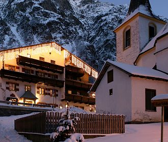 Verschneite Alpenlandschaft bei Dämmerung mit beleuchtetem Chalet und Kirche im Vordergrund, umgeben von majestätischen, schneebedeckten Berggipfeln und klarem Himmel.