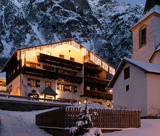 Verschneite Alpenlandschaft bei Dämmerung mit beleuchtetem Chalet und Kirche im Vordergrund, umgeben von majestätischen, schneebedeckten Berggipfeln und klarem Himmel.