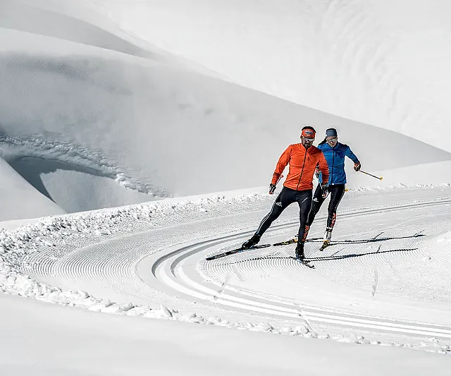 Zwei Personen skilanglaufend auf einer verschneiten Strecke, umgeben von sanften, weißen Hügeln. Der Mann trägt eine orange Jacke, die Frau eine blaue Jacke.