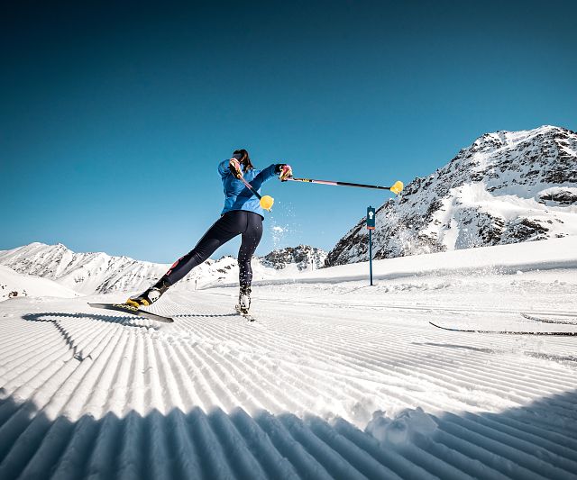 Eine Langläuferin in einer blauen Jacke, gleitet an einem hellen, klaren Tag über die Höhenloipe am Pitztaler Gletscher.