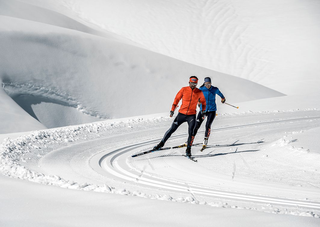 Zwei Langläufer, einer in einer orangefarbenen Jacke, der andere in einer blauen Jacke, gleiten an einem hellen, klaren Tag über präparierte Schneeloipen.