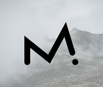 Ein stilisiertes, schwarzes „M“-Logo der Firma Maurten schwebt über einer nebligen Berglandschaft. Der Himmel ist wolkenverhangen, die Berggipfel sind teilweise verdeckt.