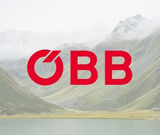 Eine malerische Landschaft mit sanften grünen Hügeln und schneebedeckten Bergen im Hintergrund. Im Vordergrund ein ruhiger See, auf dem das Logo der ÖBB in kräftigem Rot prangt.