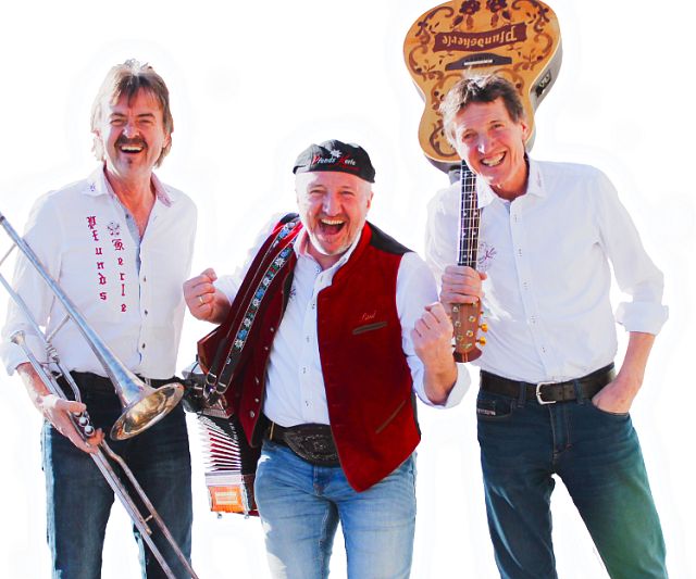 Drei Männer in weiß-roter Tracht stehen lachend nebeneinander. Einer hält eine Trompete, der andere ein Akkordeon und der dritte eine Gitarre. Alle wirken fröhlich und gut gelaunt.