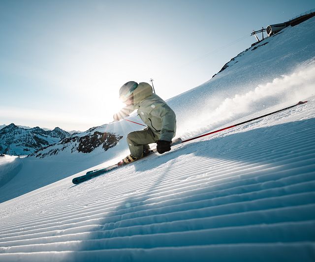 Ein Skifahrer in heller Kleidung fährt dynamisch eine frisch präparierte Piste hinab. Die Morgensonne strahlt im Hintergrund über schneebedeckte Berggipfel.
