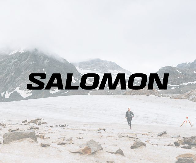 Ein Bergläufer läuft über eine schneebedeckte Landschaft mit Bergen im Hintergrund. Der Schriftzug 'SALOMON' ist prominent im Vordergrund zu sehen.
