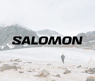 Ein Bergläufer läuft über eine schneebedeckte Landschaft mit Bergen im Hintergrund. Der Schriftzug 'SALOMON' ist prominent im Vordergrund zu sehen.