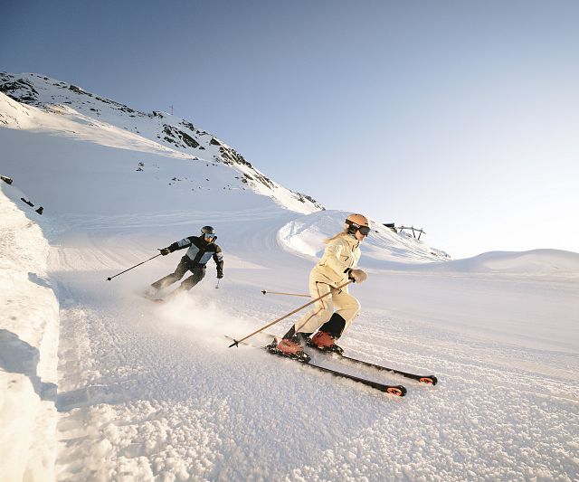 Zwei Skifahrer fahren über eine verschneite Piste in den Bergen. Der Himmel ist klar und sonnig, und die Schneedecke glänzt im Licht.