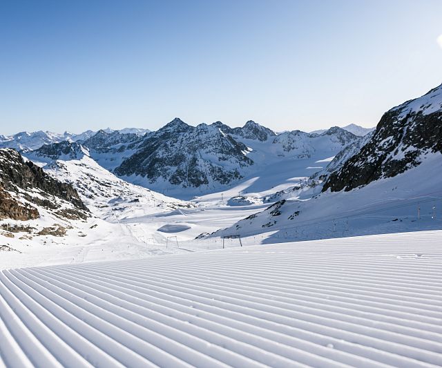 Verschneite Berglandschaft unter klarem Himmel. Im Vordergrund sind parallele Linien auf frisch präpariertem Schnee zu sehen. Die Berge erstrecken sich bis zum Horizont.