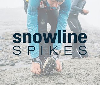 Eine Person kniet sich auf den Boden und befestigt spezielle Spikes an einem Schuh. Im Hintergrund sind verschwommene Gestalten und eine neblige Landschaft zu sehen.