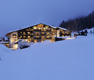 Ein beleuchtetes Hotel steht in der Abenddämmerung, umgeben von verschneiten Bäumen und Hängen, unter einem bewölkten Himmel, der eine ruhige Winterlandschaft schafft.