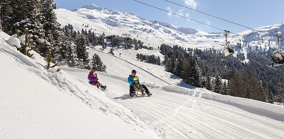 Zwei Personen mit Kindern rodeln unter einem klaren blauen Himmel die schneebedeckte Rodelbahn am Hochzeiger im Pitztal hinunter.