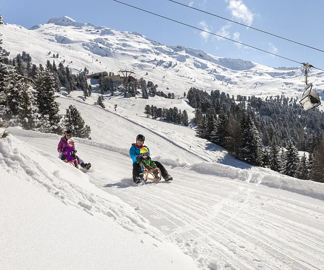 Zwei Personen mit Kindern rodeln unter einem klaren blauen Himmel die schneebedeckte Rodelbahn am Hochzeiger im Pitztal hinunter.