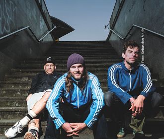 Drei Männer in blauen Jacken sitzen auf einer Treppe im Freien. Einer trägt eine Mütze, die anderen sehen entspannt aus. Die Treppe ist von grauen Wänden umgeben.