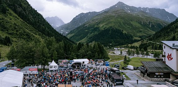 Luftaufnahme eines belebten Outdoor-Events in einem alpinen Tal mit Bergen im Hintergrund. Menschen versammeln sich um Stände und eine Bühne, umgeben von grüner.
