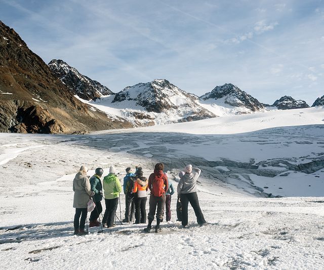 Eine Gruppe von Menschen steht in winterlicher Kleidung auf Schnee im Vordergrund. Im Hintergrund sind schneebedeckte Berge unter einem klaren Himmel zu sehen.