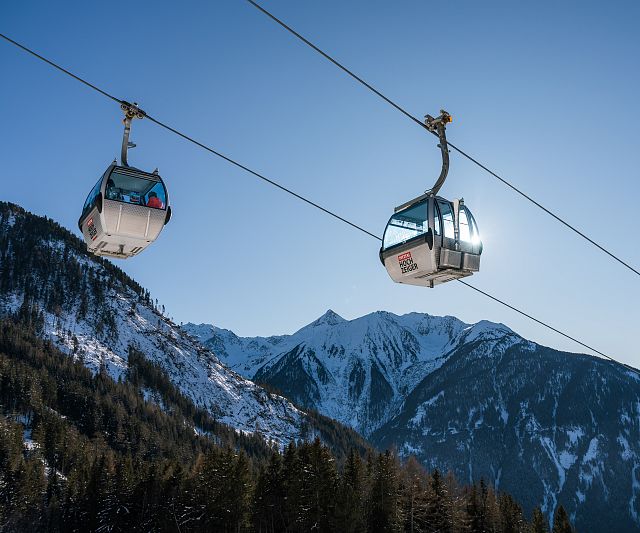 Zwei Gondeln schweben an Kabeln über einer schneebedeckten Berglandschaft. Im Hintergrund sind beeindruckende, sonnendurchflutete Berggipfel unter einem klaren blauen Himmel zu sehen.