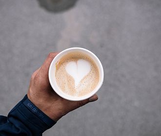 Eine Nahaufnahme einer Hand, die einen Becher mit Latte Macchiato hält. Der Milchschaum hat die Form eines Herzens, vor einem grauen, unregelmäßigen Untergrund.