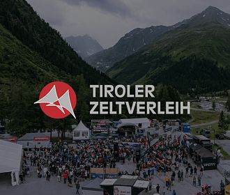 Panoramablick auf eine große Menschenmenge bei einem Veranstaltungsort in den Bergen. Im Vordergrund sind Zeltstände und eine Bühne zu sehen.